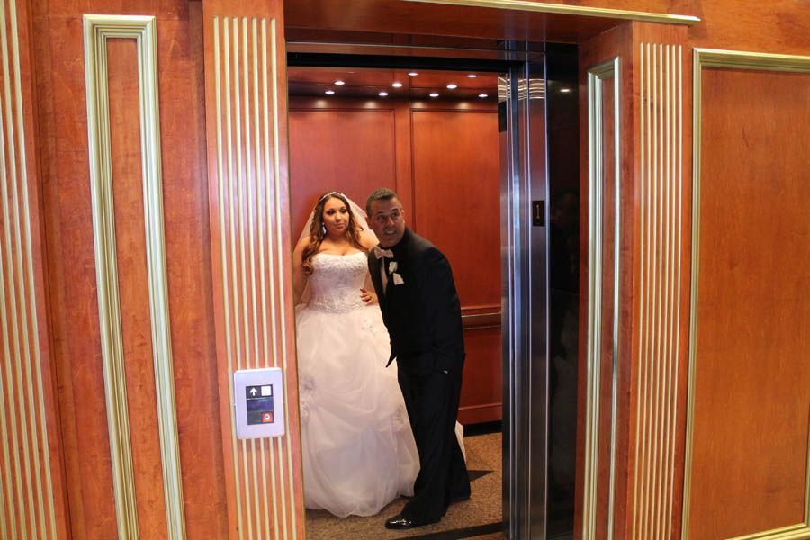 Bride in a wedding gown and groom in a tuxedo exiting a wood-paneled elevator.