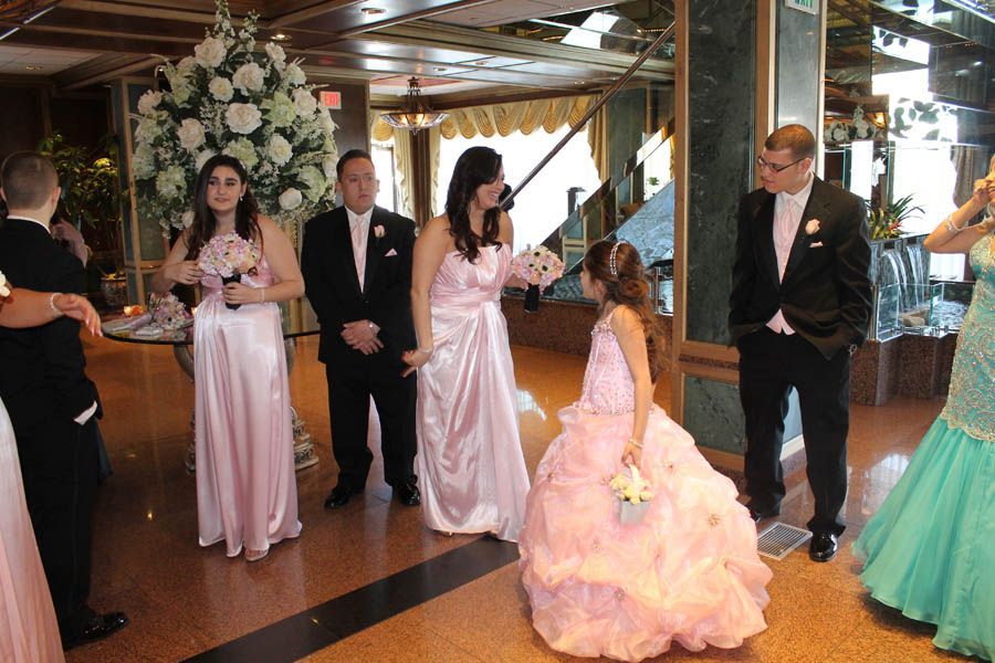 Wedding party in formal attire poses indoors; bridesmaids in pink gowns, groomsmen in black suits, flower girl in pink dress.