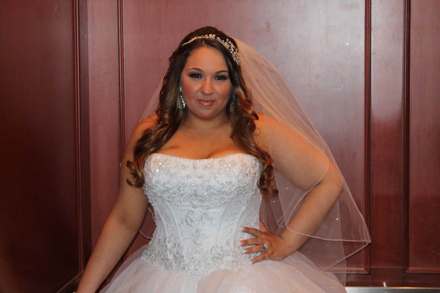 Bride in a white strapless gown with a veil, posing with a hand on her hip against a wood panel wall.