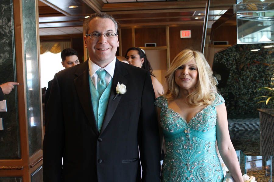 Couple smiling, the man in a black suit, teal tie, and the woman in a teal dress, walking indoors.