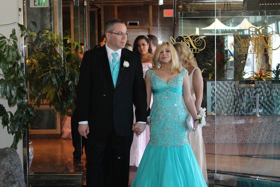 Couple in formal attire, holding hands, entering a building, with bridesmaids in background. Teal and black.