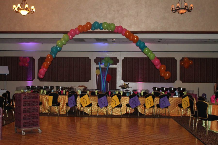 Balloon arch over a decorated banquet table with gold tablecloths and colored chair covers.