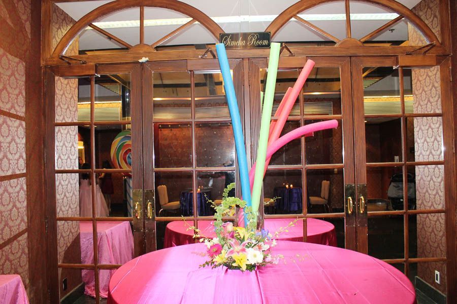 Event table with pink tablecloth, floral arrangement, and colorful inflatable tubes in front of wooden double doors.