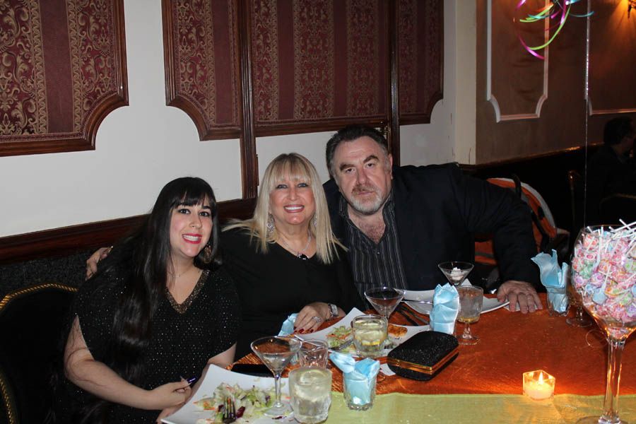 Three people smiling at a table set for dinner. Orange tablecloth, food, and drinks visible.