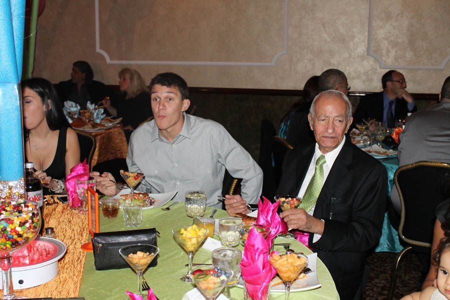People seated at a festively decorated table, enjoying a meal. Some look at the camera. Green tablecloth, colorful food.