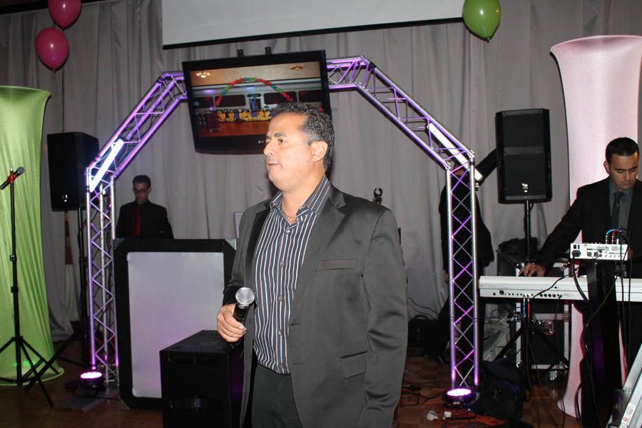 Man with microphone speaks at event, stage setup with lights, screen, and keyboard players.
