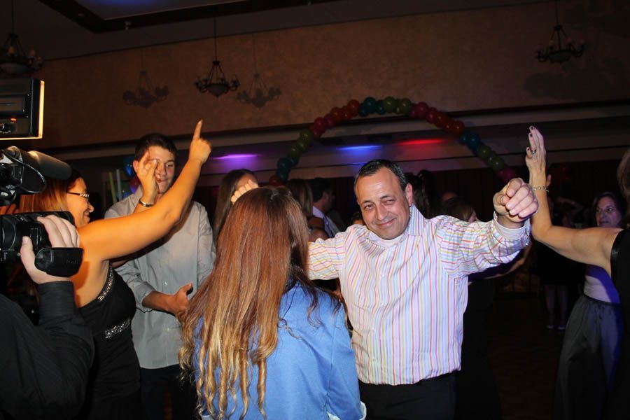 People dancing at a party under a colorful balloon arch. Man in striped shirt dances with arms raised.