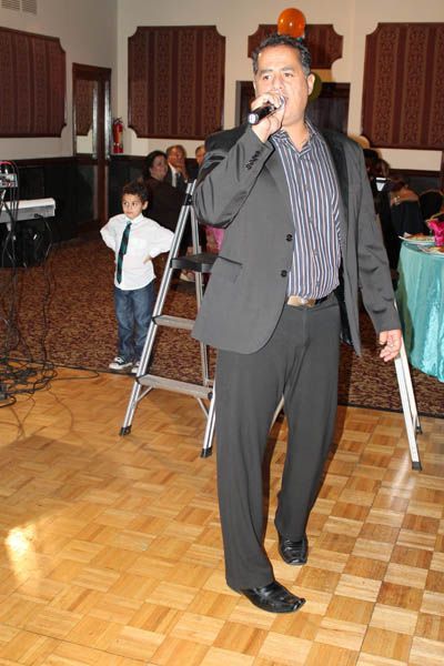 Man singing into microphone. Young boy standing on a stepladder in an event hall with wooden floors.