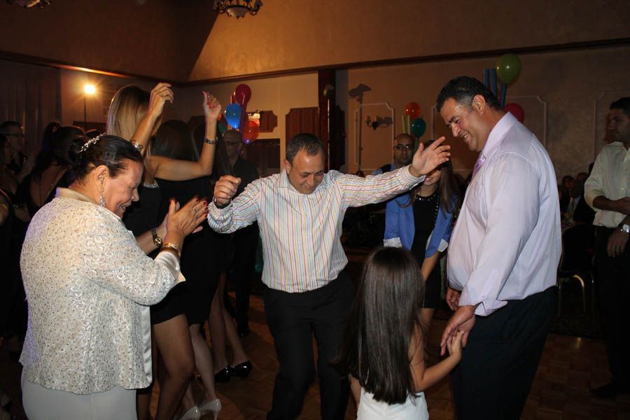 People dancing in a well-lit room; a man dances with arms outstretched, another man smiles, and a child dances nearby.