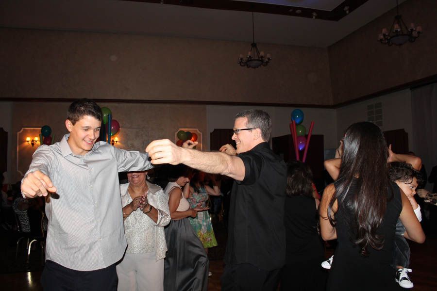 People dancing at a party in a large hall with balloons and chandeliers; man in black shirt and teenager in gray shirt dance.