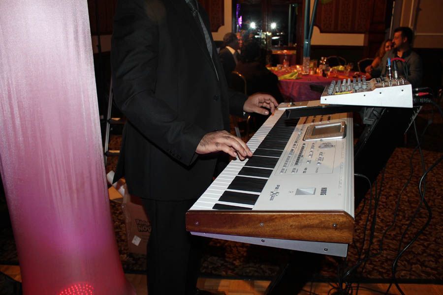 Person in black suit playing a keyboard on a stand; event in background.