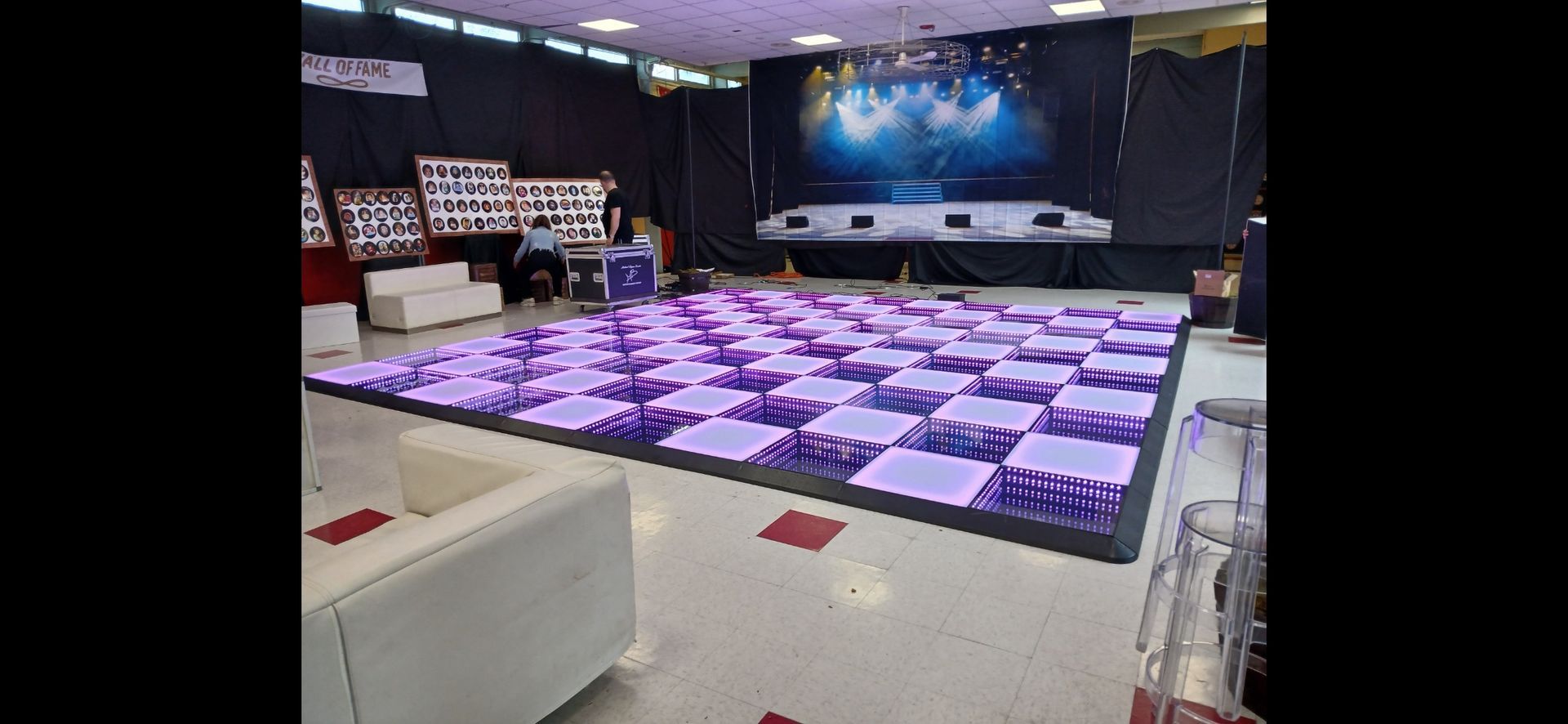 Lit-up checkered dance floor in a venue. Stage lights in the background. White sofas and people seated in the middle.