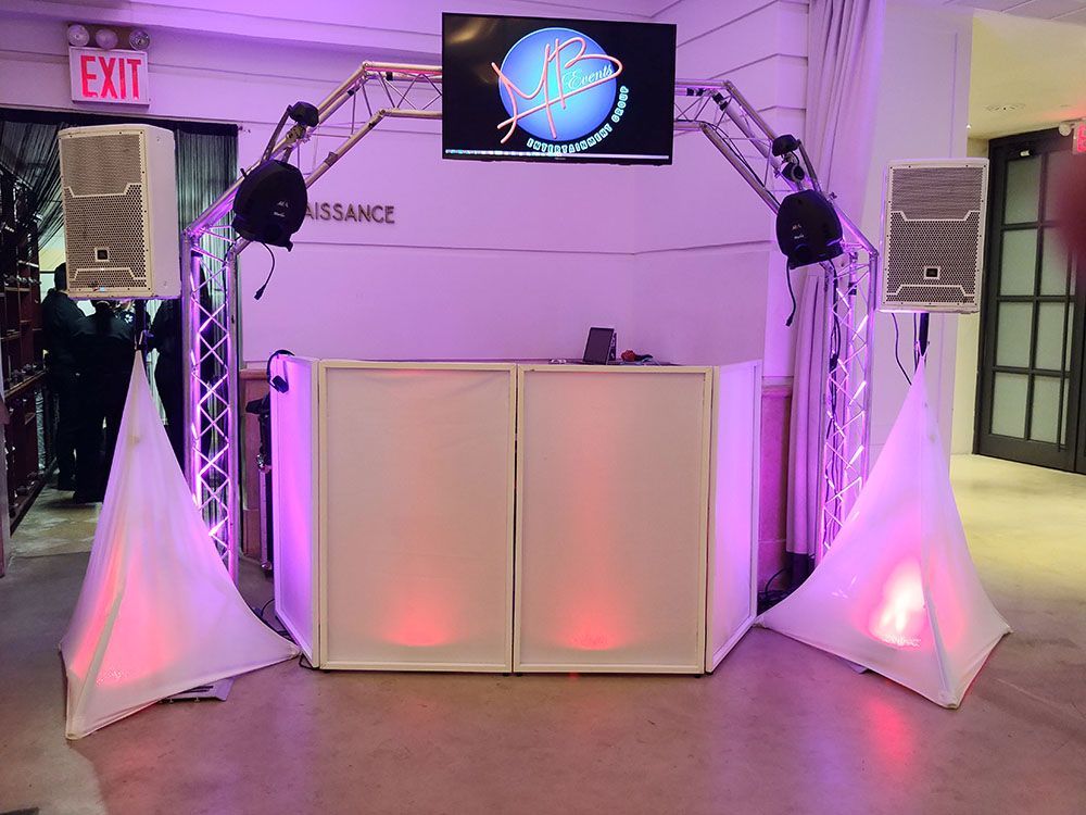 DJ setup with speakers, truss, white facade, screen, and uplighting. Interior with exit sign.