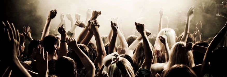 People with raised arms in a concert crowd, illuminated by stage lights.