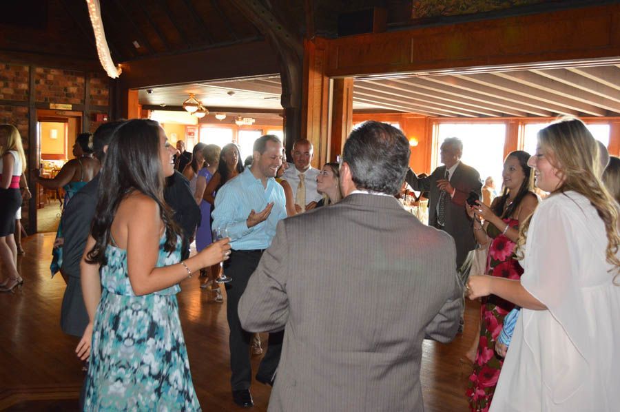 People dancing and socializing at a reception in a wood-paneled room with natural light.