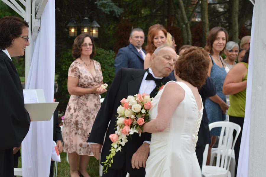 Couple embraces after a wedding ceremony under a white arbor, guests watching.