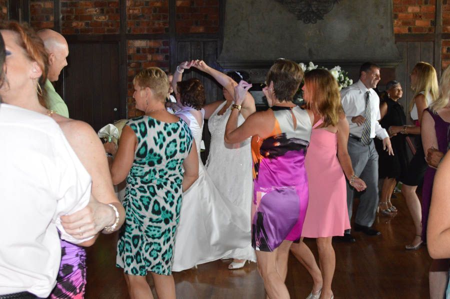 People dancing at a wedding reception. Bride in white dress, guests in colorful dresses, brick wall background.