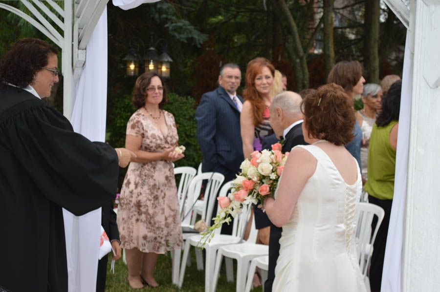 Wedding ceremony: bride, groom, officiant, guests, decorated archway, outdoor setting.