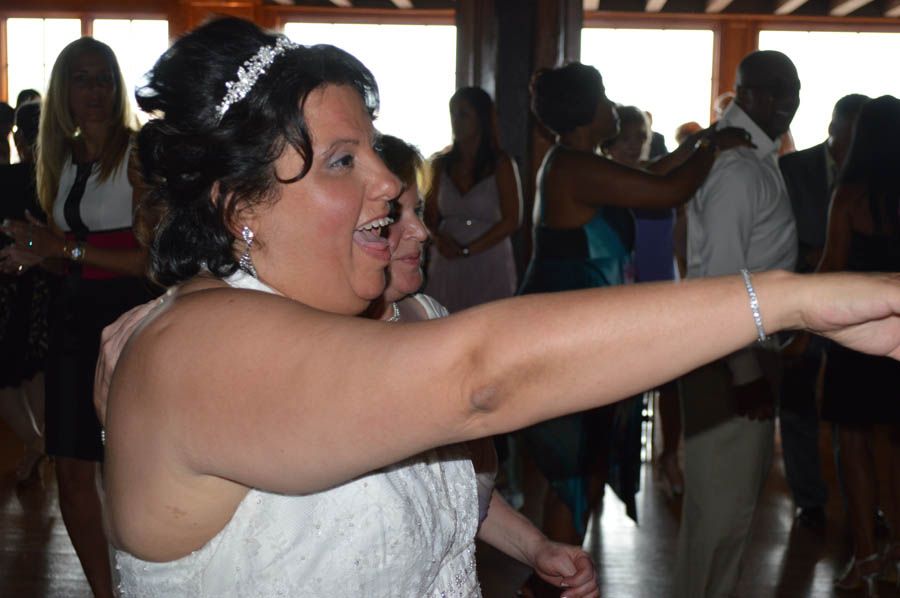 Bride dancing, arm extended, smiling, wearing a white dress. People in background.