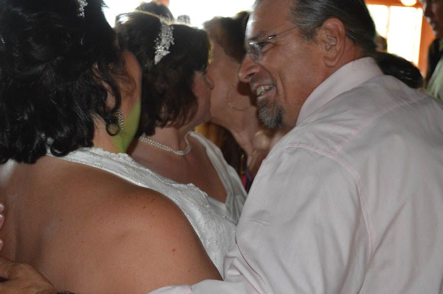 Wedding guests embracing, smiling. Bride in white dress, man in pink shirt.