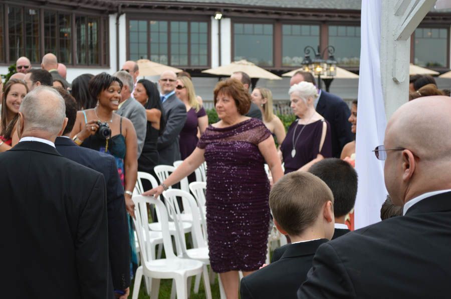 People at an outdoor wedding ceremony; woman in purple dress moves chairs.