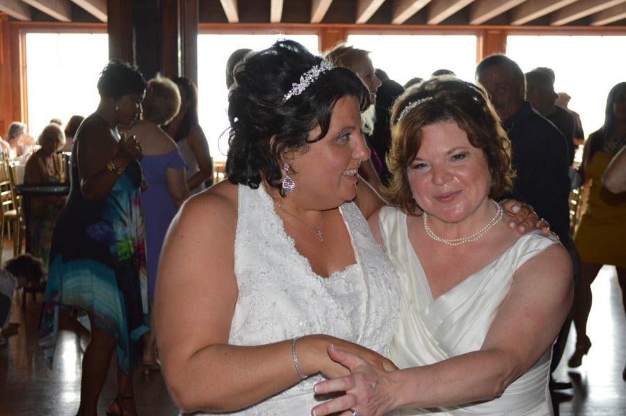 Two women in white dresses embrace at a celebration. Soft light and people in background.
