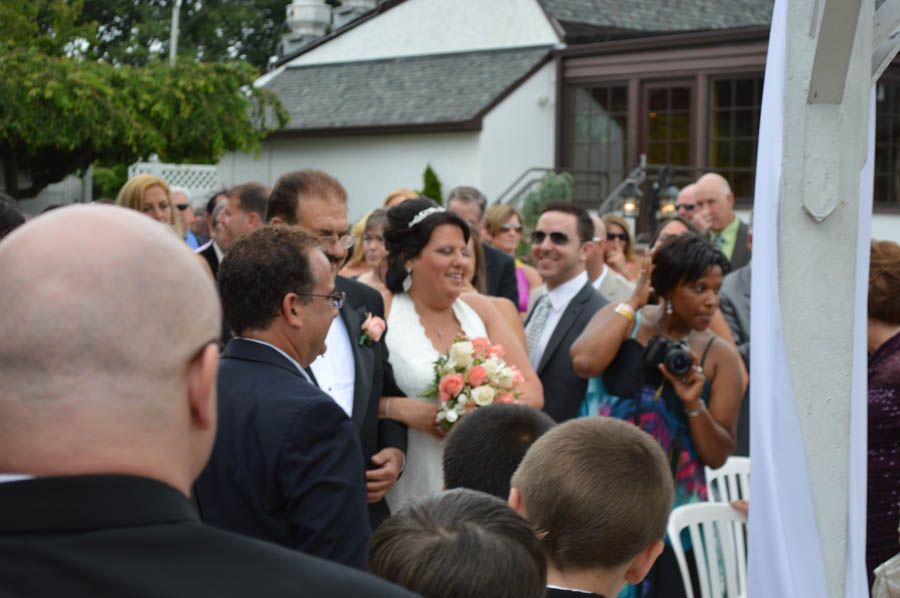 Bride smiling, walking down aisle at outdoor wedding with guests in the background.