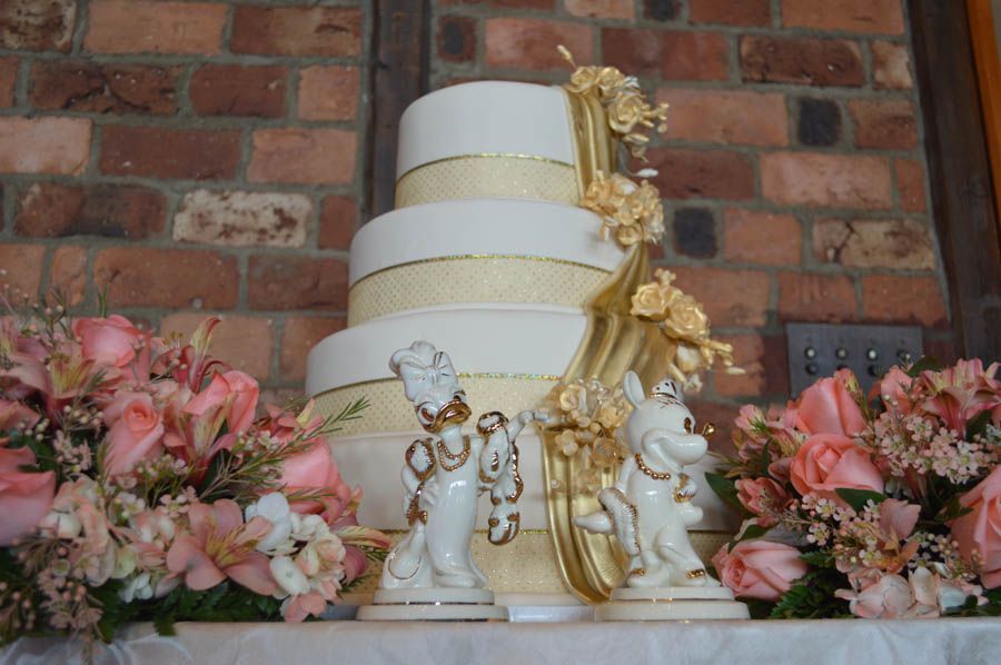 Wedding cake, three tiers, gold ribbons, flanked by pink flowers and figurines, against a brick wall.