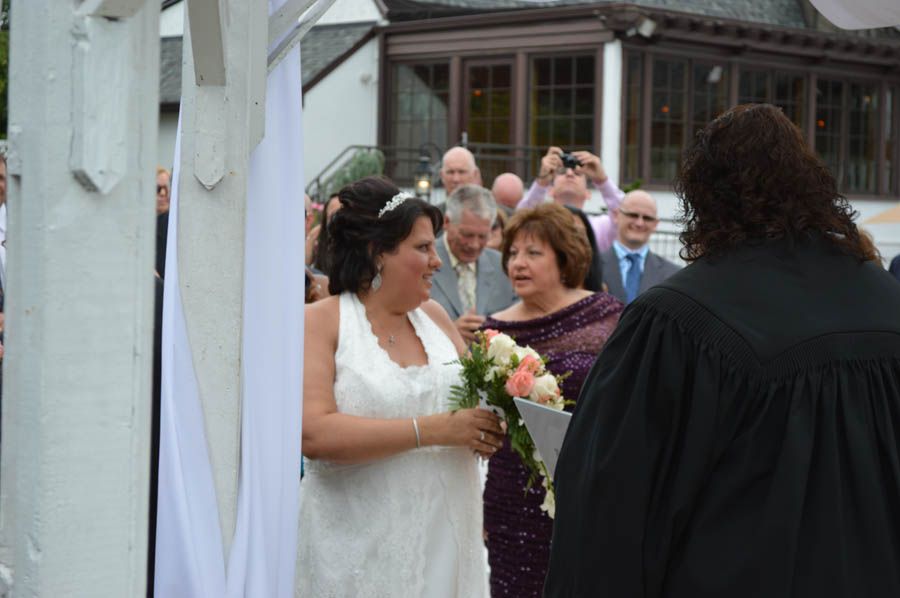 Bride smiles during a wedding ceremony outdoors, holding flowers. Other guests watch in the background.