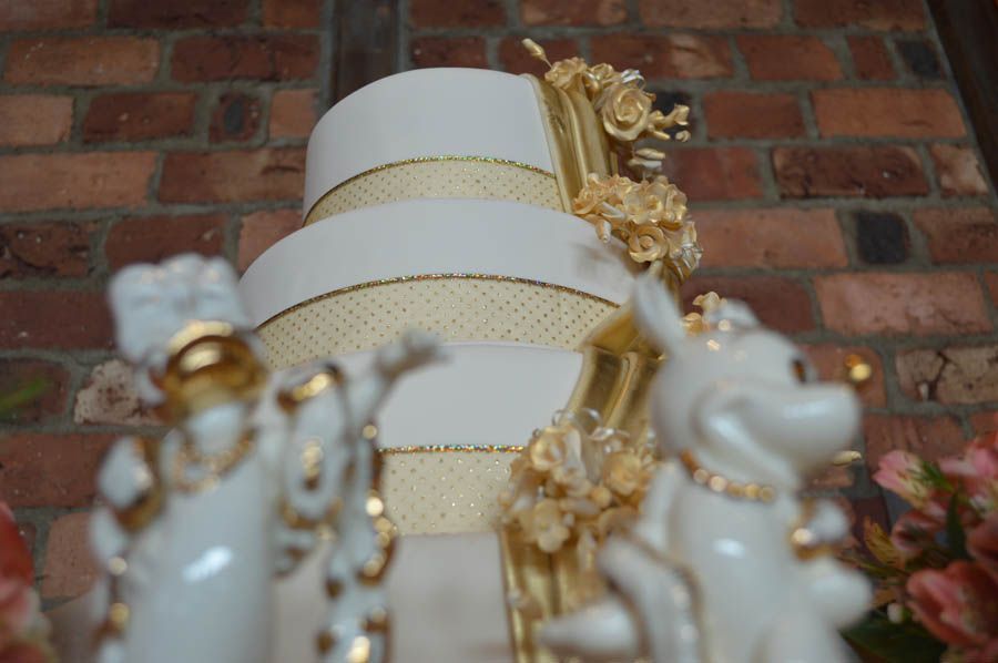 Three-tiered white wedding cake with gold ribbon and floral decorations, figurines in foreground, brick background.