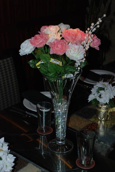 Flowers in a clear vase with water beads on a dark table, glasses, and a smaller flower arrangement.