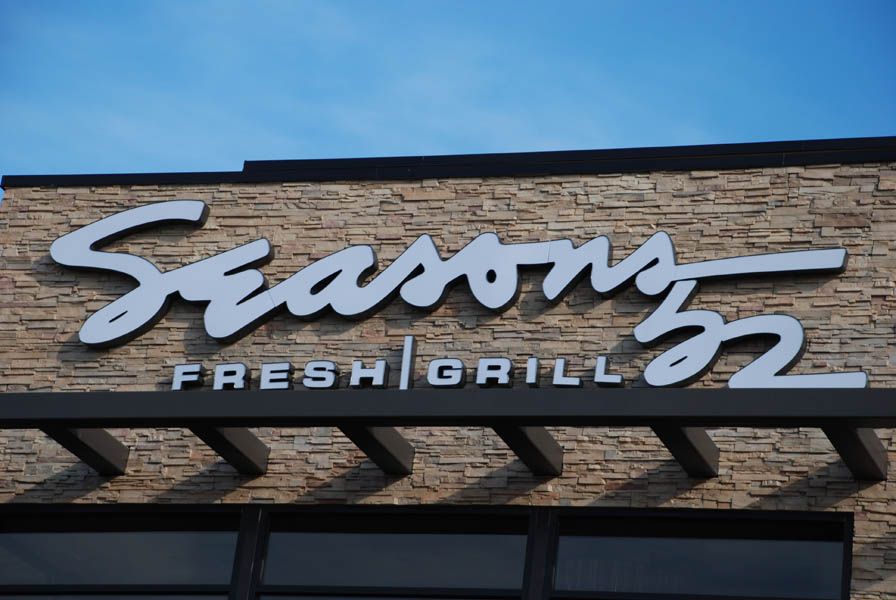 Sign for Seasons 52 Fresh Grill restaurant; silver lettering on a stone facade under a blue sky.