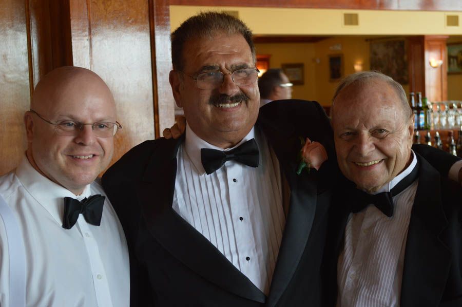 Three men in tuxedos, smiling at the camera, inside a restaurant with wooden paneling.