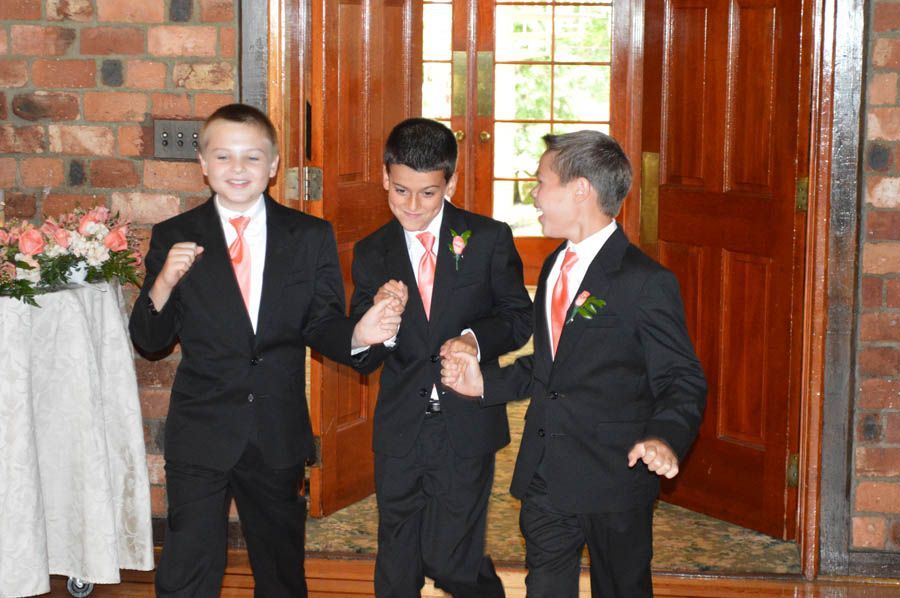 Three boys in black suits and coral ties, entering a building, smiling, arms linked.