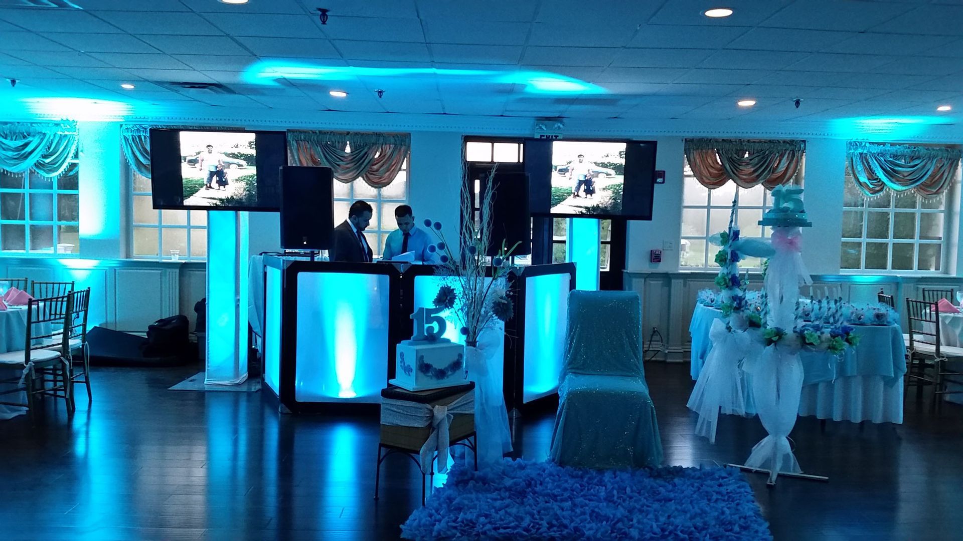A DJ setup at an event, lit with blue lights. Two people are behind the equipment.