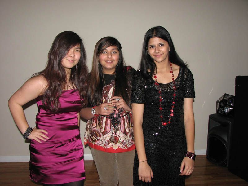 Three young women pose indoors; one in a pink dress, one in a patterned top, and one in a black sequined dress.