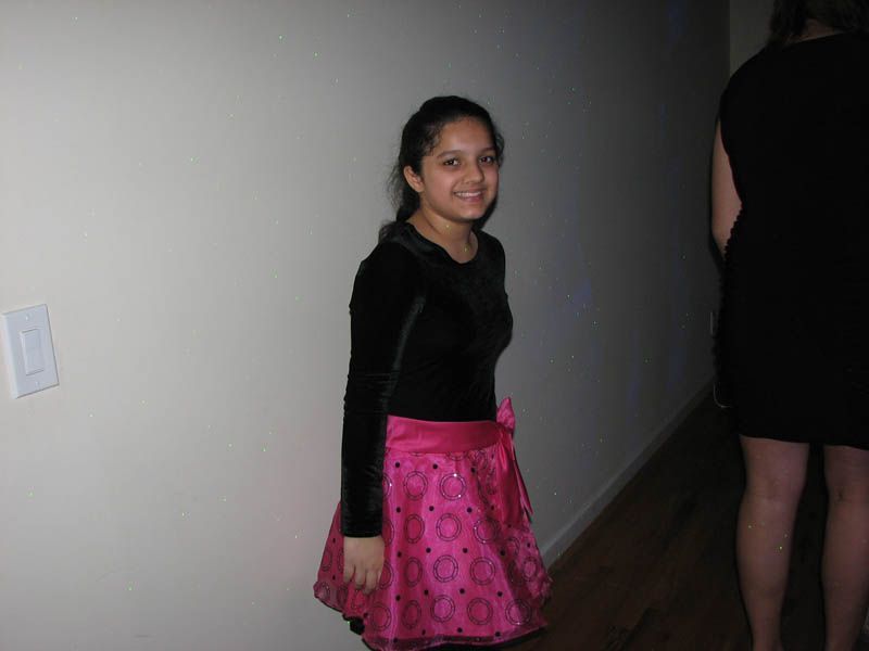 Person in black top, pink skirt smiles near a wall. Another person in black dress on the right.