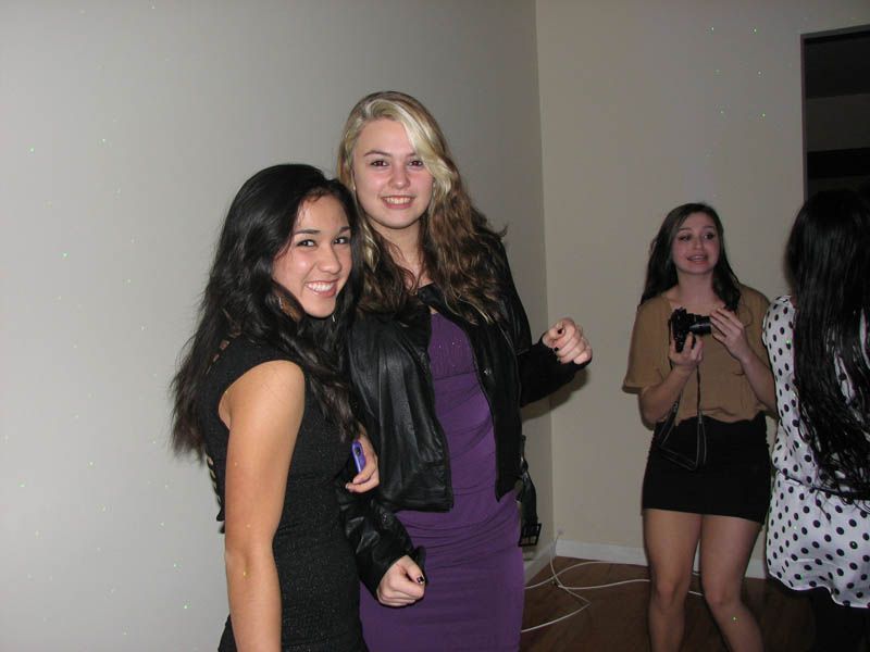 Two women smiling and posing at a party, one in a black dress, the other in a purple dress and leather jacket.