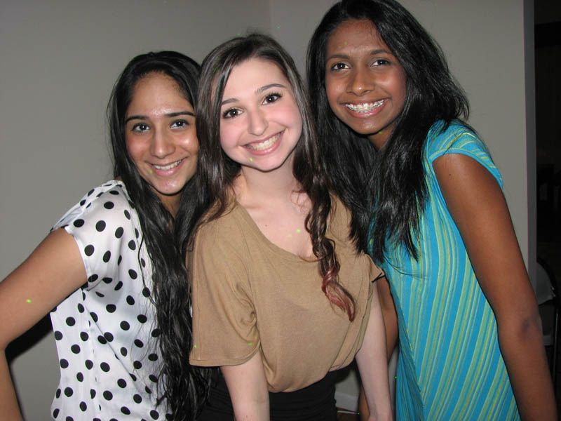 Three smiling women posing closely together indoors; one in polka dots, one in tan, one in blue.