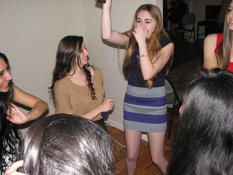 Group of young people dancing in a room; woman holding object overhead and covering mouth.