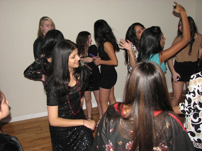 A group of young people gathered indoors, some dancing and interacting, against a neutral wall.