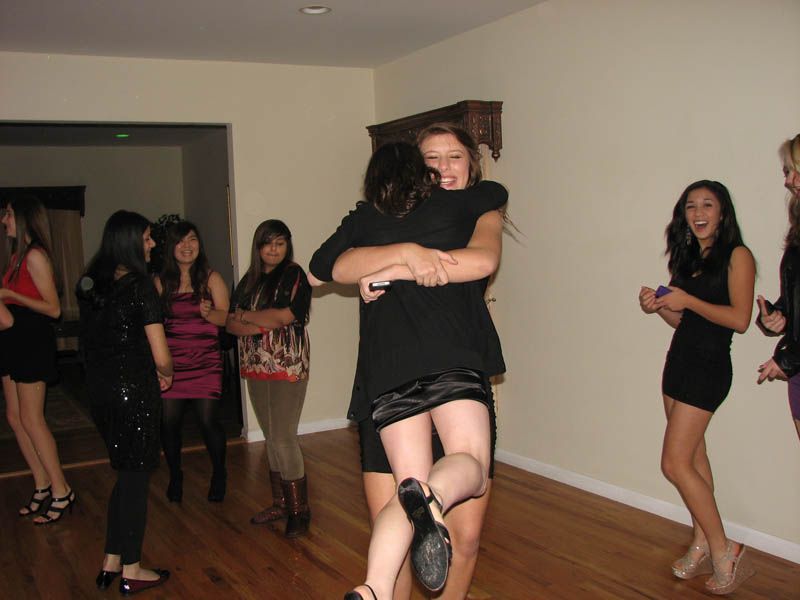 Women hugging at a party in a well-lit room with wooden floors. Others watch and smile.