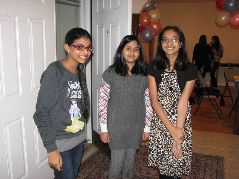 Three girls in front of an open door; one in a patterned dress, others in sweaters. Balloons and people in the background.