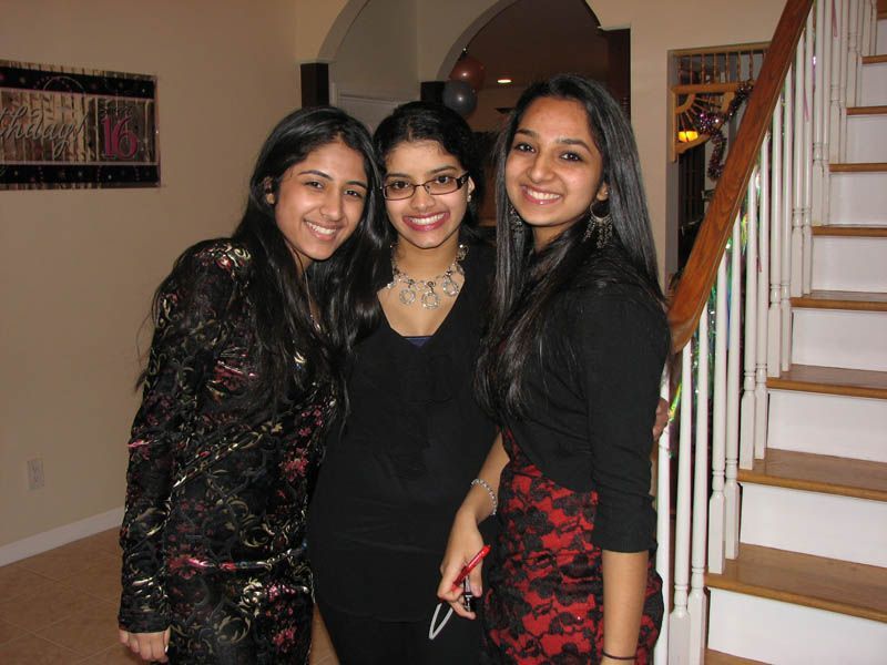 Three young women smiling, posing for a photo indoors near a staircase, one in a black dress with red lace.