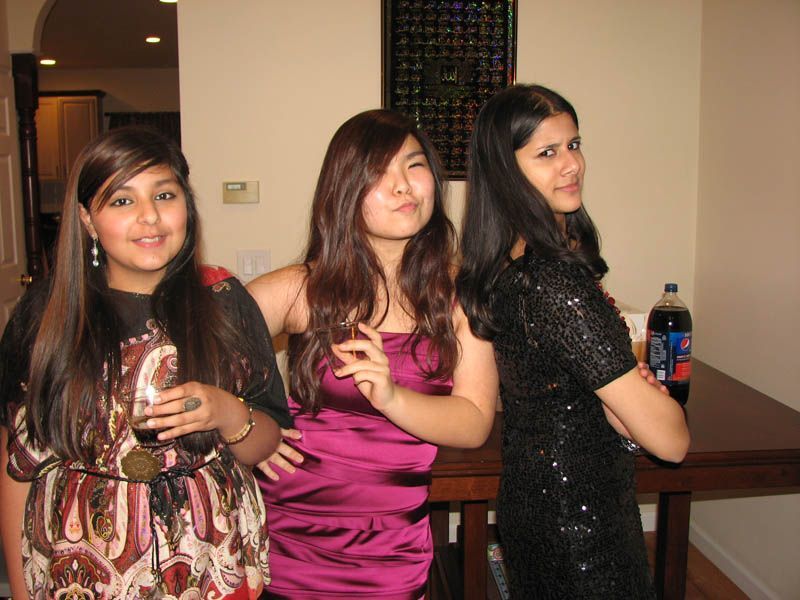 Three young women posing indoors; one with drink, one smirking, and one with arms crossed, next to table.