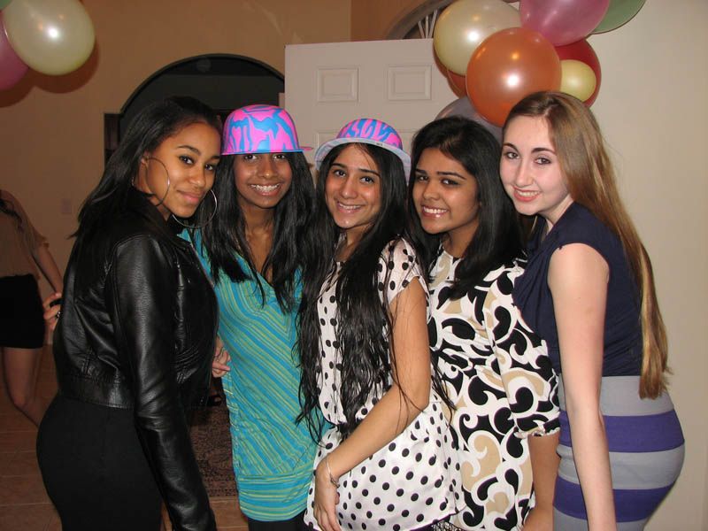 Five young people pose together at a party, wearing casual clothes and hats, with balloons in the background.