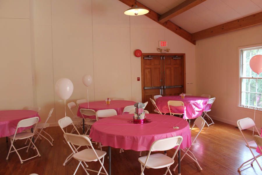 Room with round tables covered in pink tablecloths, white chairs, and balloons; a light hangs from the ceiling.