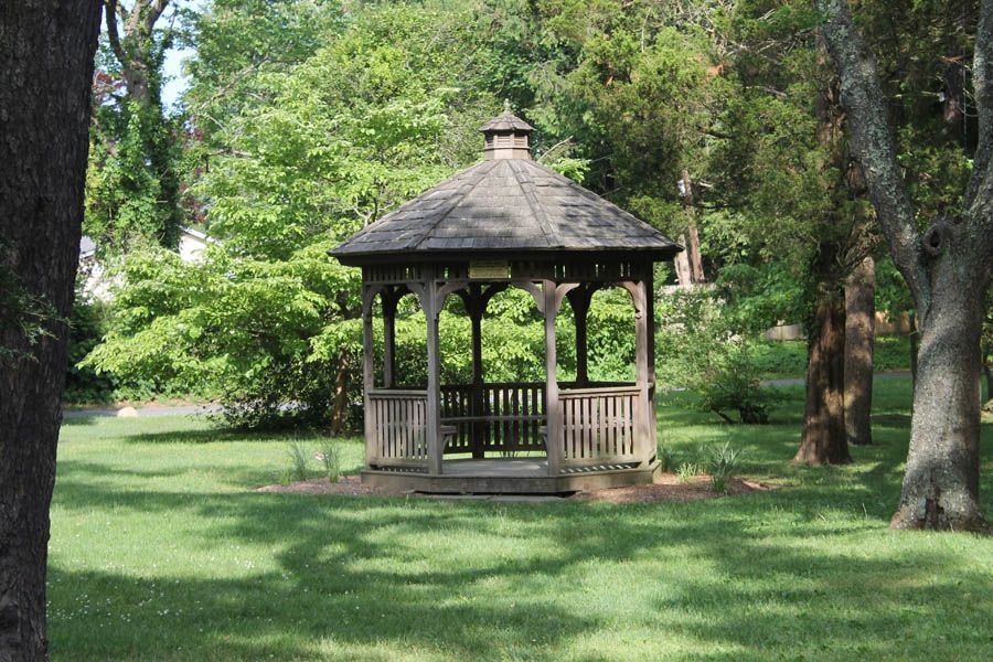 Wooden gazebo with a peaked roof in a grassy park, surrounded by trees and dappled sunlight.