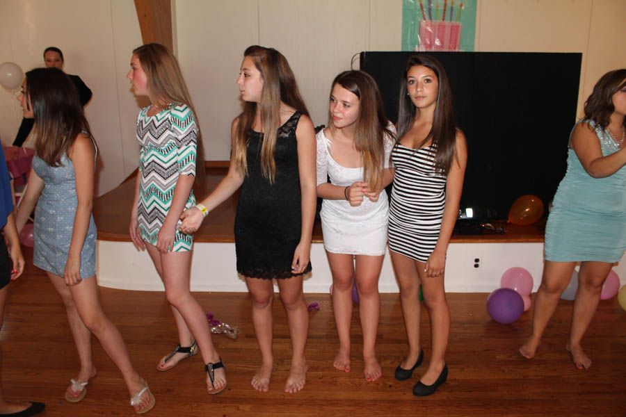 Several young people stand on a wood floor, dressed in party attire. Some are barefoot.