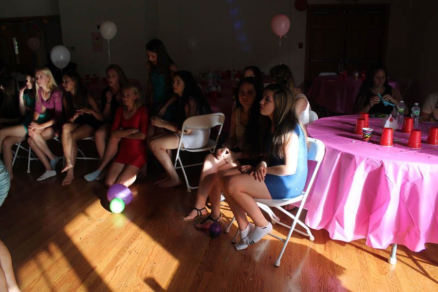 People at a party, seated in chairs, observing a table with pink tablecloth and drinks.
